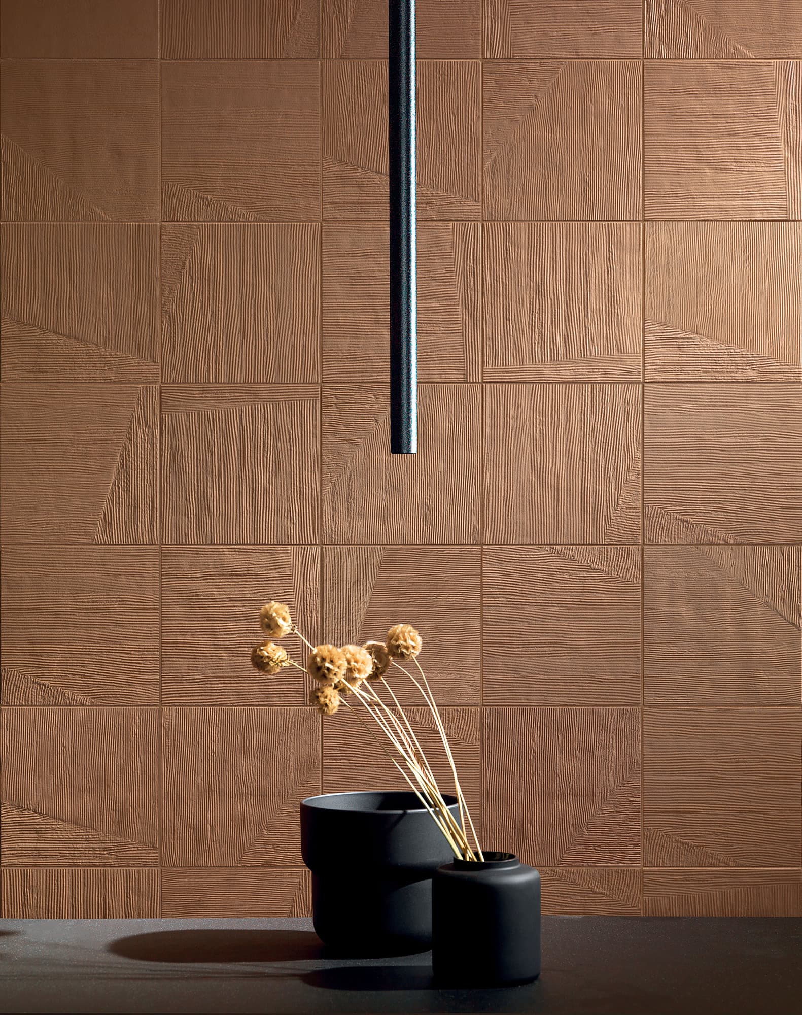 Detaljbilde interiør med flislagt cottofarget vegg med tekstur. Foran veggen synes et sort bord med en sort vase med blomster.